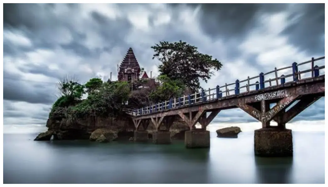 Ambil foto Pura Ismoyo dari kejauhan saat senja, menangkap siluet dramatisnya dengan langit jingga.