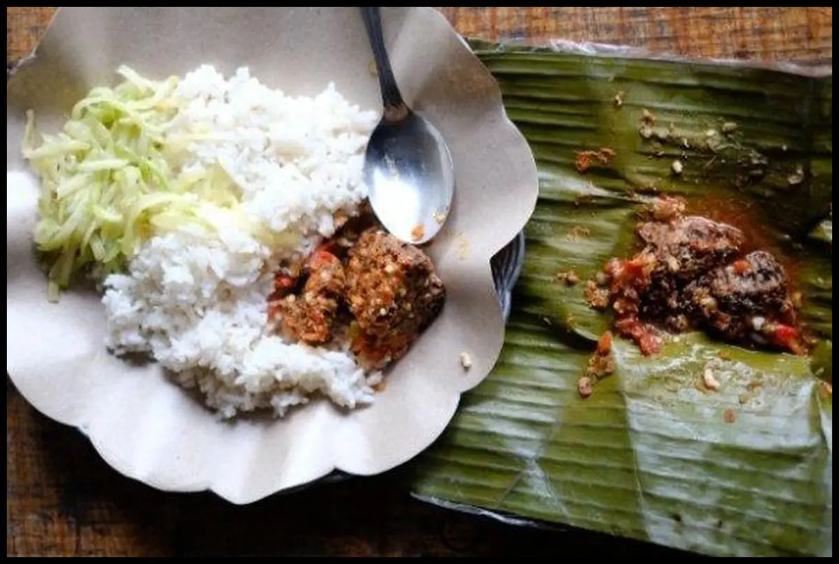 Sudut pandang mata burung (top-down) yang memperlihatkan tumpukan lauk pauk di atas pincuk daun pisang dengan latar belakang gerbang Stasiun Kotabaru.