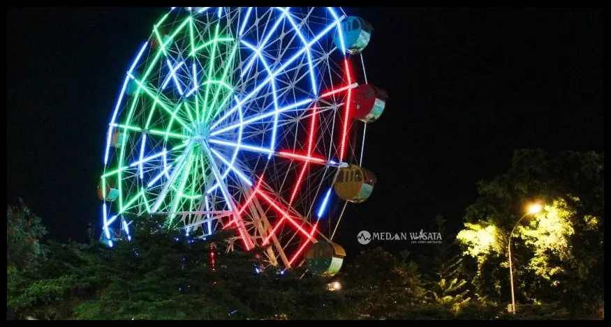 Pose sambil memegang piring ketan dengan latar belakang lampu bianglala yang menyala warna-warni sekitar pukul 19.00 WIB.