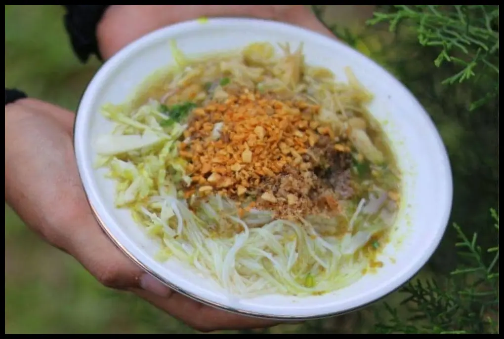 Close-up kepulan asap dari mangkuk soto yang baru saja disiram kuah panas, fokus pada taburan koya kuning yang perlahan meleleh di atas nasi.