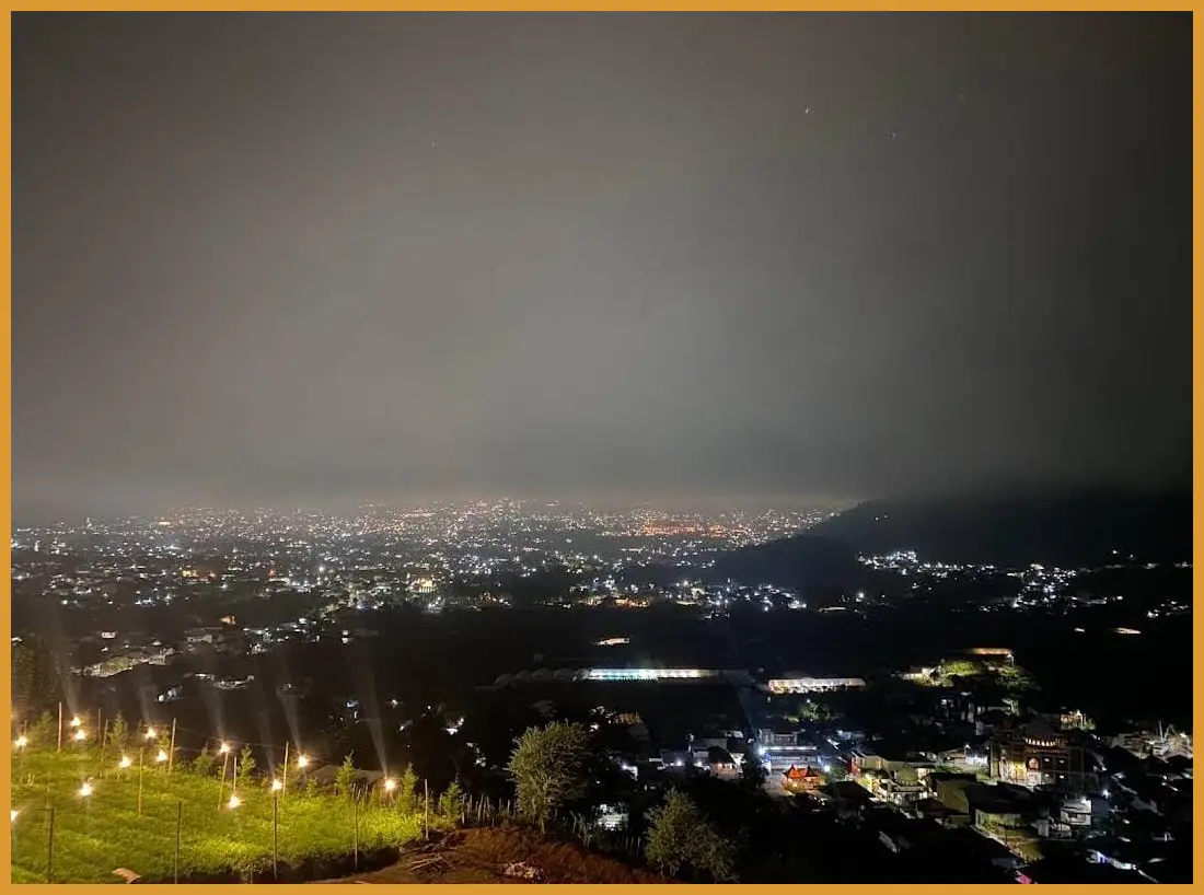Foto dari arah jalan raya yang menangkap kemegahan fasad bangunan kafe dengan latar belakang lampu kendaraan yang berlalu-lalang (light trail).